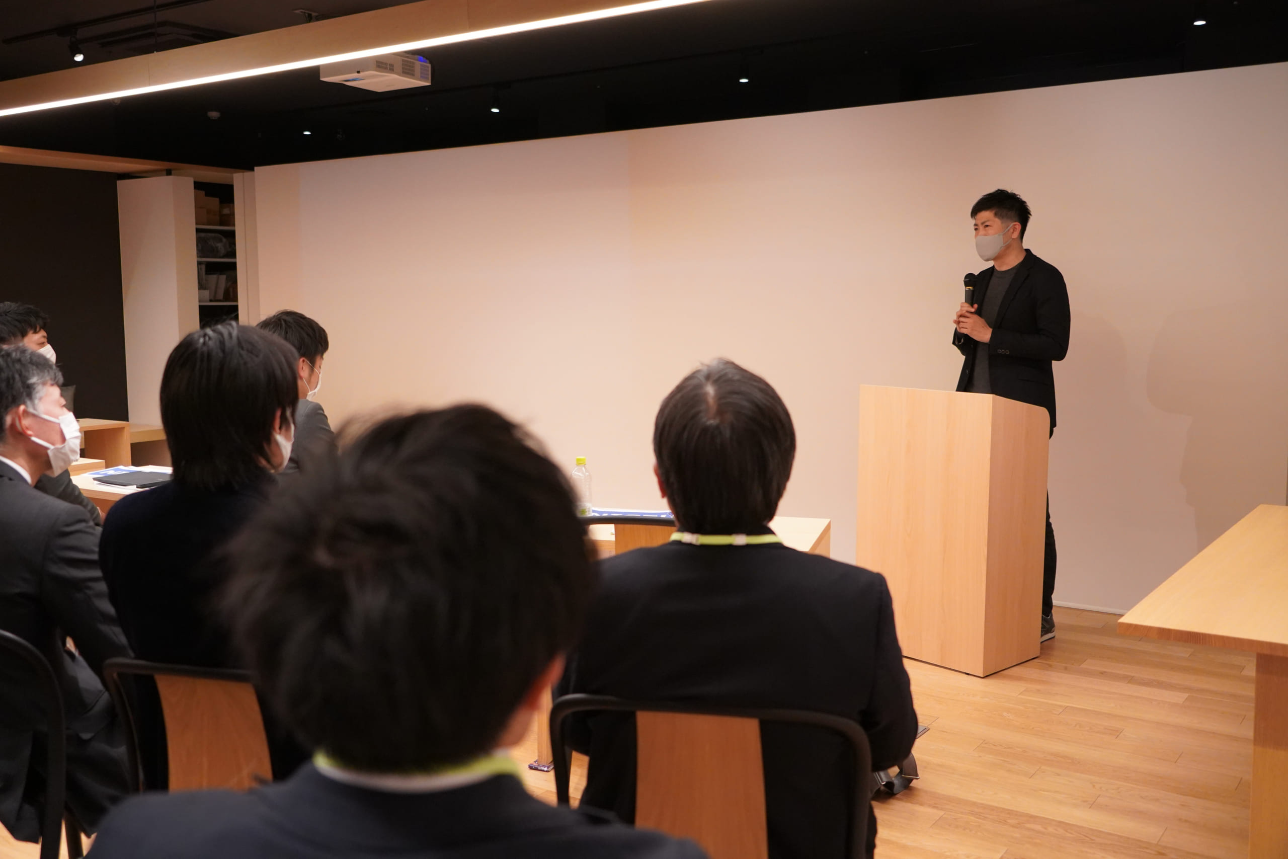 MZV's CEO Makino is standing behind the speaker podium while a bunch of attendees listening to him